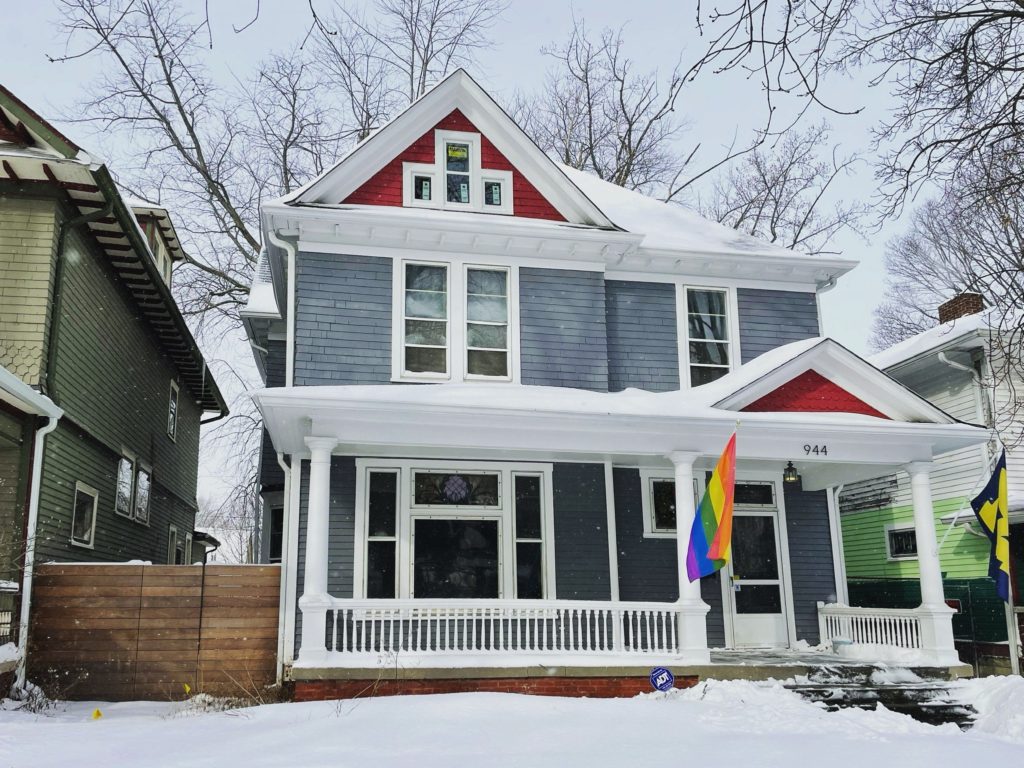 A Queen Anne style house after renovation. The house is blue and red with white trim and a rainbow LGBT flag in front. Snow gently blankets the house. It has two stories and a porch.
