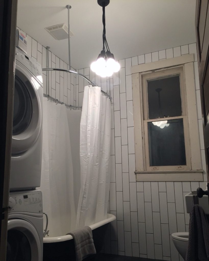The Queen Anne bathroom after renovation. It has white vertical subway tile walls and a washer/dryer now, but it retains the antique clawfoot tub. There is a new shower curtain, plain white.
