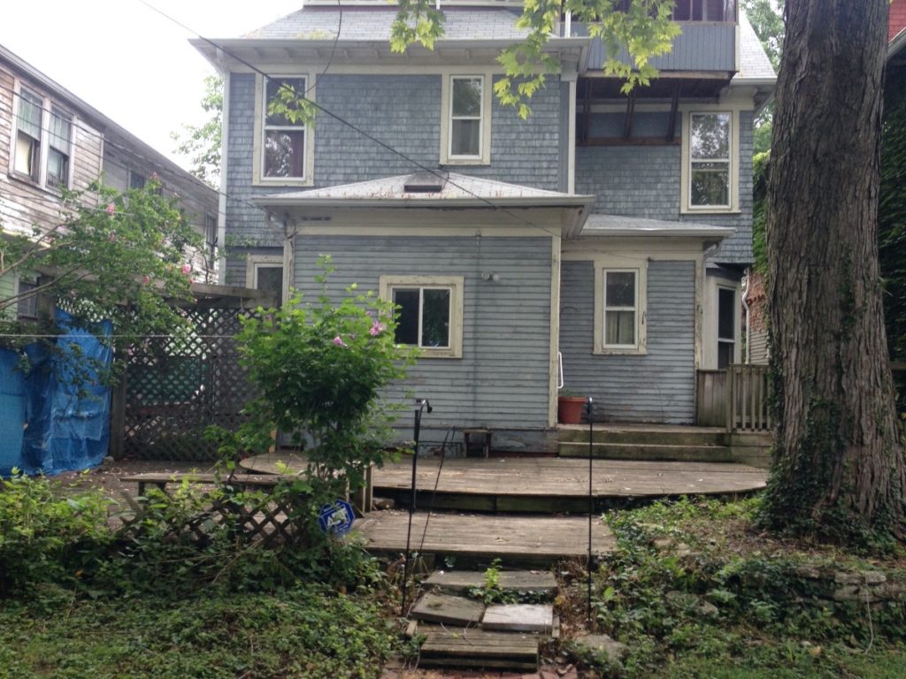 A before picture of the back yard renovation. The yard is overgrown. The siding looks worn and needs a paint job, and the patio is uneven and aged.