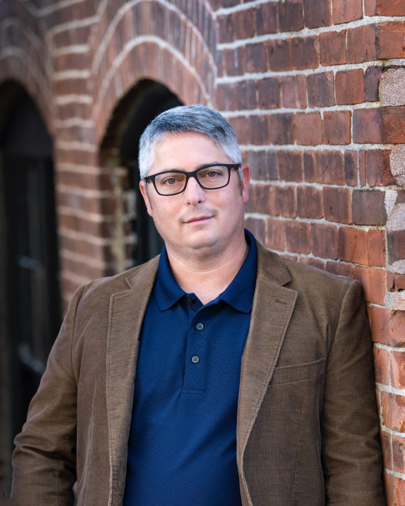 A medium close-up of architect Chris Novelli standing against a brick wall. He has light skin, short gray hair, and rectangular glasses, and is wearing a navy polo shirt and tan, professorial sport jacket.