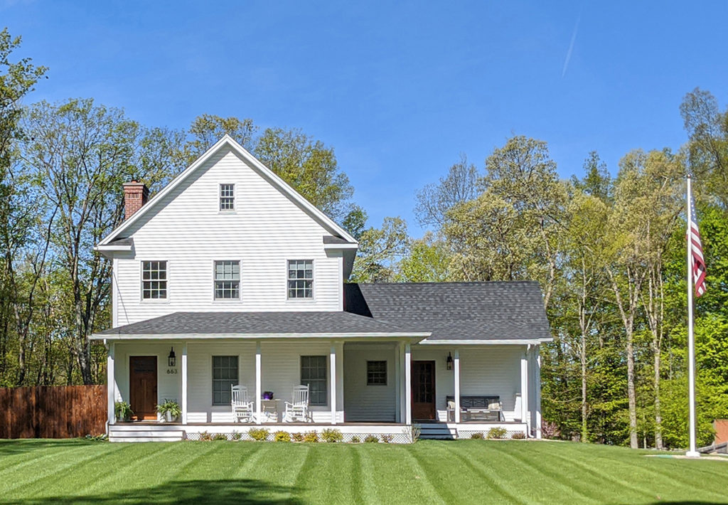 A photo of a white two-story farmhouse with a front porch on a well-manicured lawn. An American flag hangs from a pole on the right.