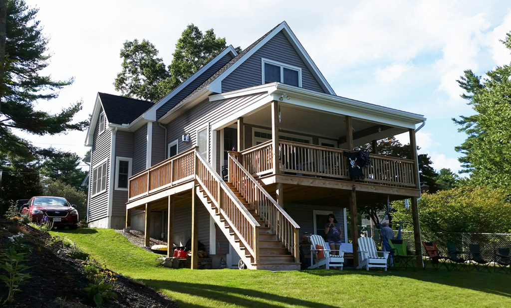 A photo of the back of a two-story house with a big deck. Stairs lead down to the lawn, where two chairs and several people sit.