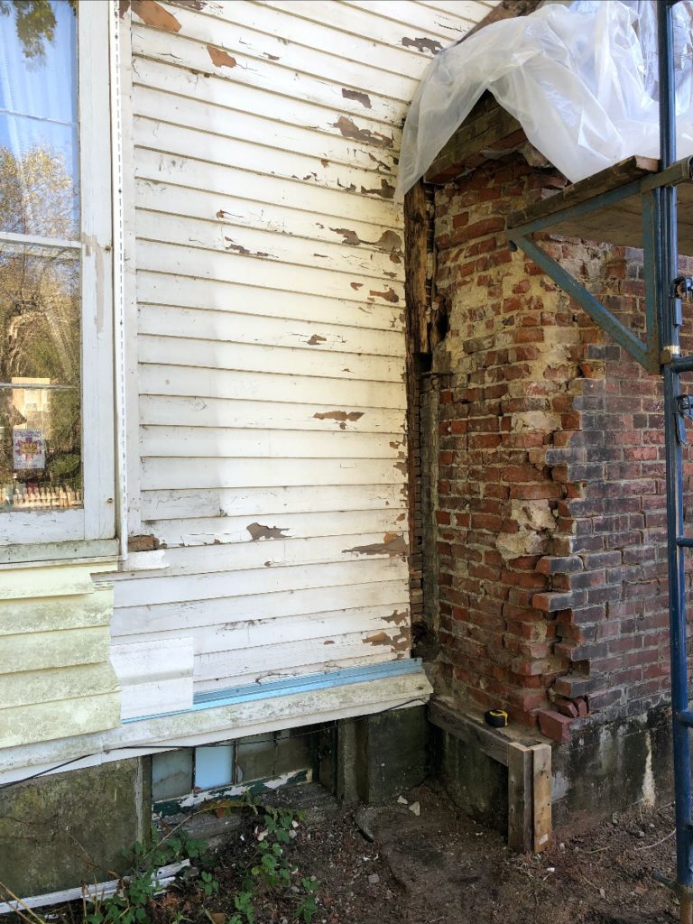 An exterior wall of a house. Vinyl siding has been removed, showing peeling old wood siding underneath.