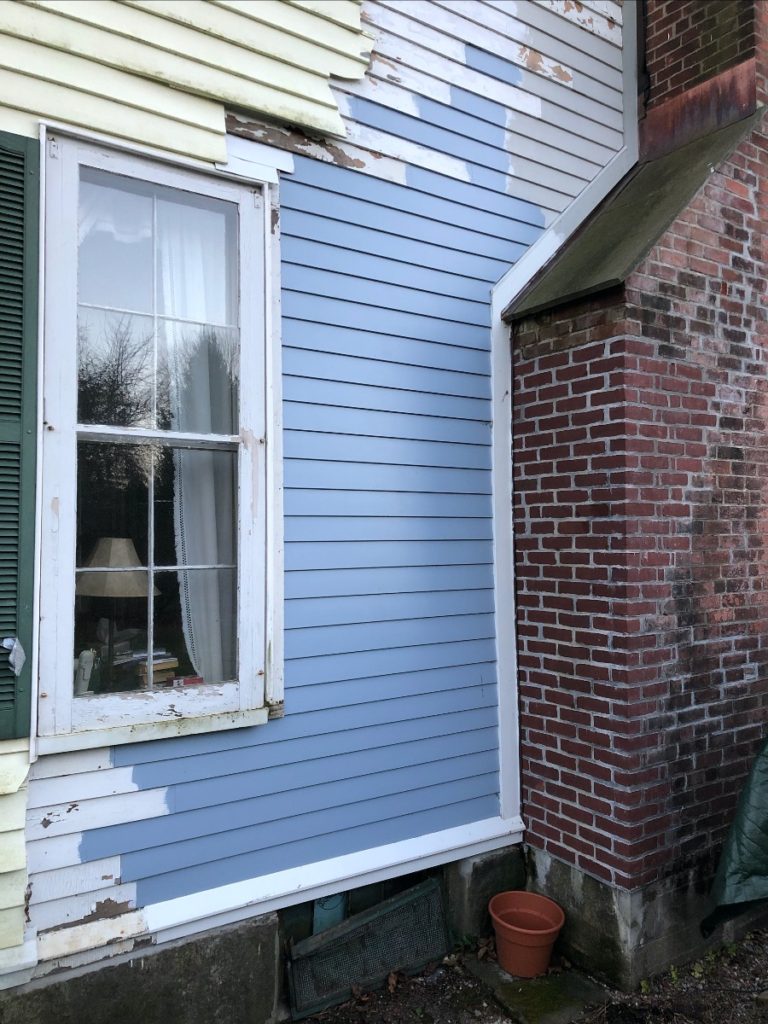 The same exterior wall of a house. The peeling paint has been cleared and the old wood siding has been painted gray and looks much nicer.